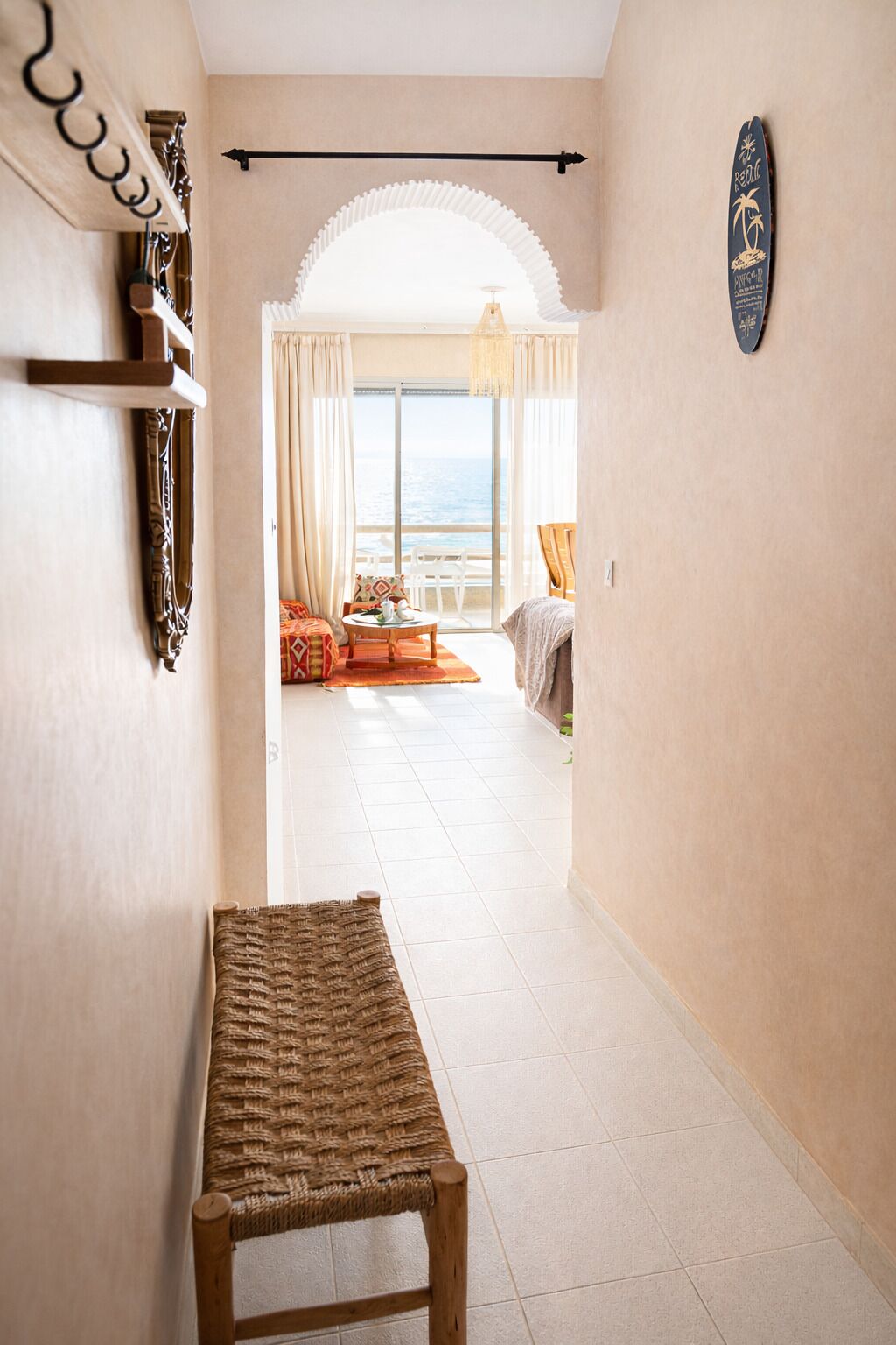 Un couloir menant à une pièce offrant une vue sur la mer, avec un banc en bois et une étagère fixée au mur.