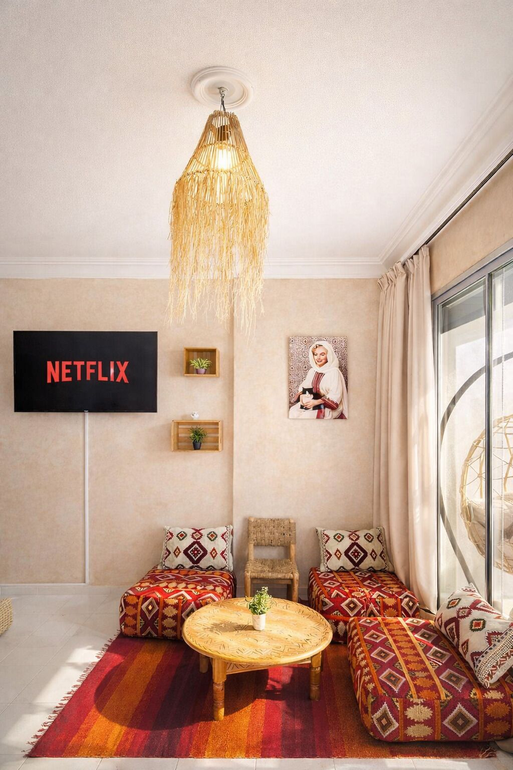 A cozy living room with a large window, a hanging light fixture, a wall-mounted TV, and patterned cushions.