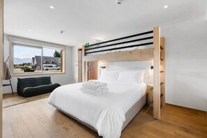 Une chambre moderne avec un lit superposé, une grande fenêtre offrant une belle vue, une table de chevet et un canapé de couleur sombre.
