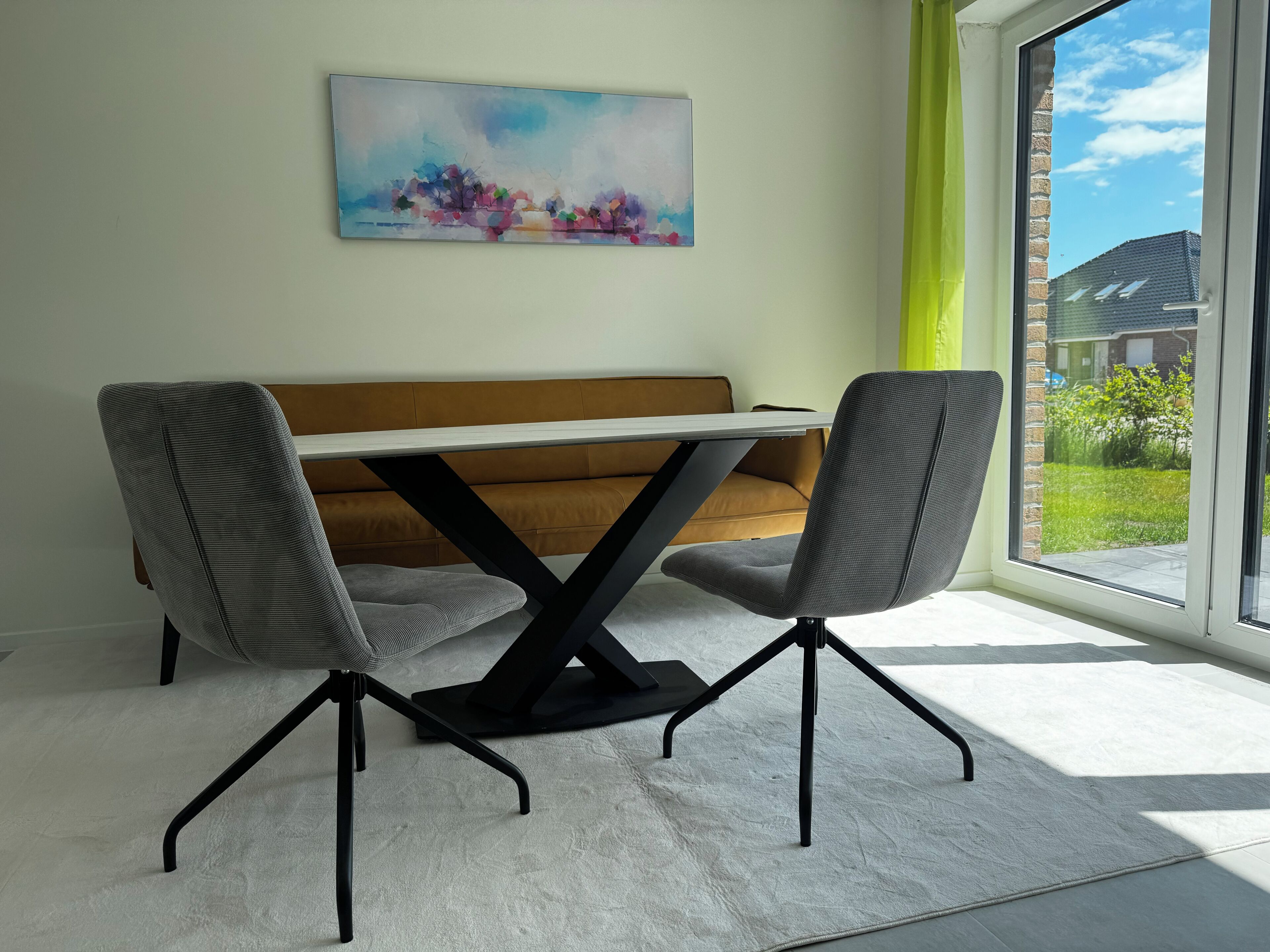 A modern dining area with a white table, two grey chairs, a brown sofa, and a large window with a view of a garden.