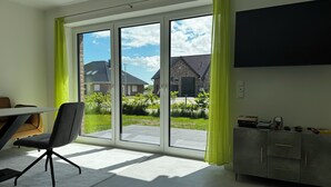 Una habitación con puerta de vidrio corrediza, una mesa de comedor con sillas, un sofá gris, un televisor montado en la pared y un mueble gris con cajones.