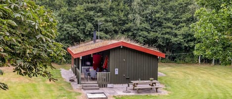A small wooden cabin with a red roof, a covered patio area, and a picnic table.