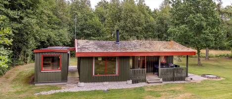 Une petite cabane dotée d’une véranda et d’une remise séparée, le tout entouré d’arbres.