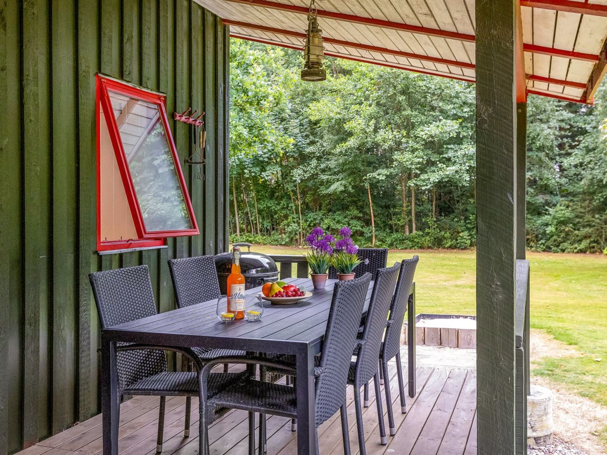 A covered outdoor dining area with a table and chairs, a grill, and a view of the surrounding greenery.