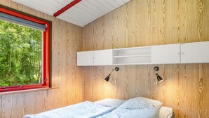 Ein Schlafzimmer mit Bett, Fenster mit rotem Rahmen, Holzvertäfelung und einem weißen Kopfteil mit Regalen.