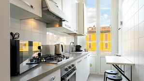 Cozinha moderna com fogão a gás, forno e máquina de café, uma mesa pequena com banquetas e vista para edifícios amarelos pela janela.