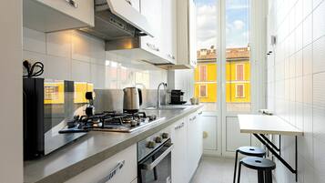 Una cucina moderna con piano cottura a gas, forno e macchina del caffè, un piccolo tavolo con sgabelli e vista su edifici gialli attraverso una finestra.
