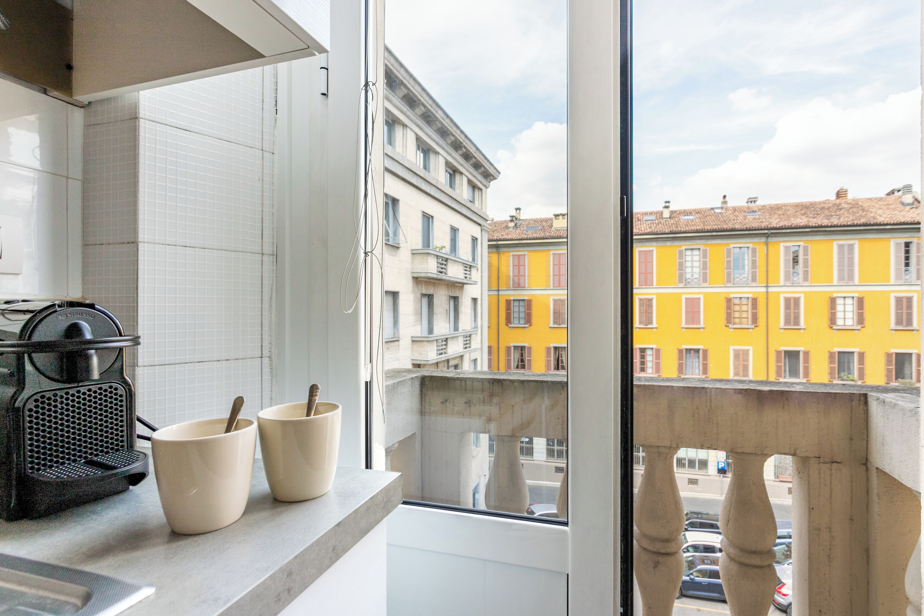 Una cucina con una macchina per il caffè, due tazze e vista su edifici colorati attraverso una finestra.