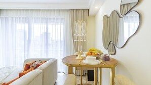 Una camera d'albergo con un letto, un angolo salotto, un tavolo da pranzo con frutta e un vaso, e uno specchio decorativo.
