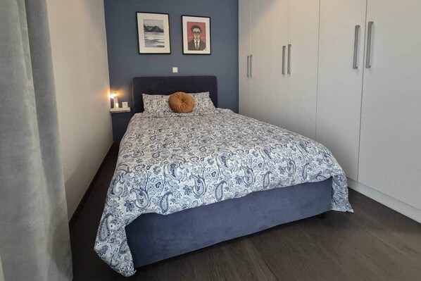 A bedroom with a bed, a nightstand, a lamp, a wardrobe, and two framed pictures on the wall.