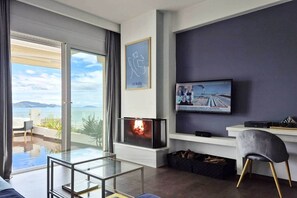 A modern living room with a TV, a fireplace, a glass table, and a view of the sea.