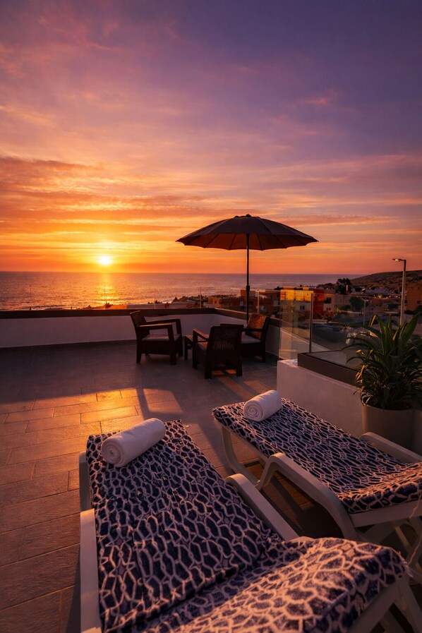Terraza en la azotea con sillones de descanso, una mesa y una sombrilla, con vista a la puesta de sol.