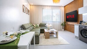 A modern living room with a sectional sofa, a dining table set for two, a coffee table, a TV with 'NETFLIX' on it, a washing machine, and a large window with sheer curtains. - Couple Haven•Workspace & WiFi 200Mb/s•Free Parking (Imi Ouaddar)