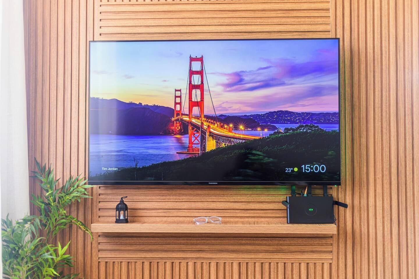 Een flatscreen-tv bevestigd aan een houten wand, met een landschapsafbeelding van een brug, een kleine plant en een decoratief object eronder.