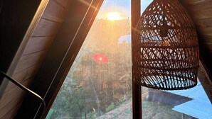 A room with a sloped ceiling, a woven light fixture, and a view of a forested area through a window.
