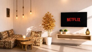A modern living room with a sofa, a coffee table, hanging lights, a potted plant, a TV displaying 'NETFLIX,' and a shelf with decorative items. - Luxury Berber Chic•3mn to Beach•Rooftop•Balcony•AC (Imi Ouaddar)