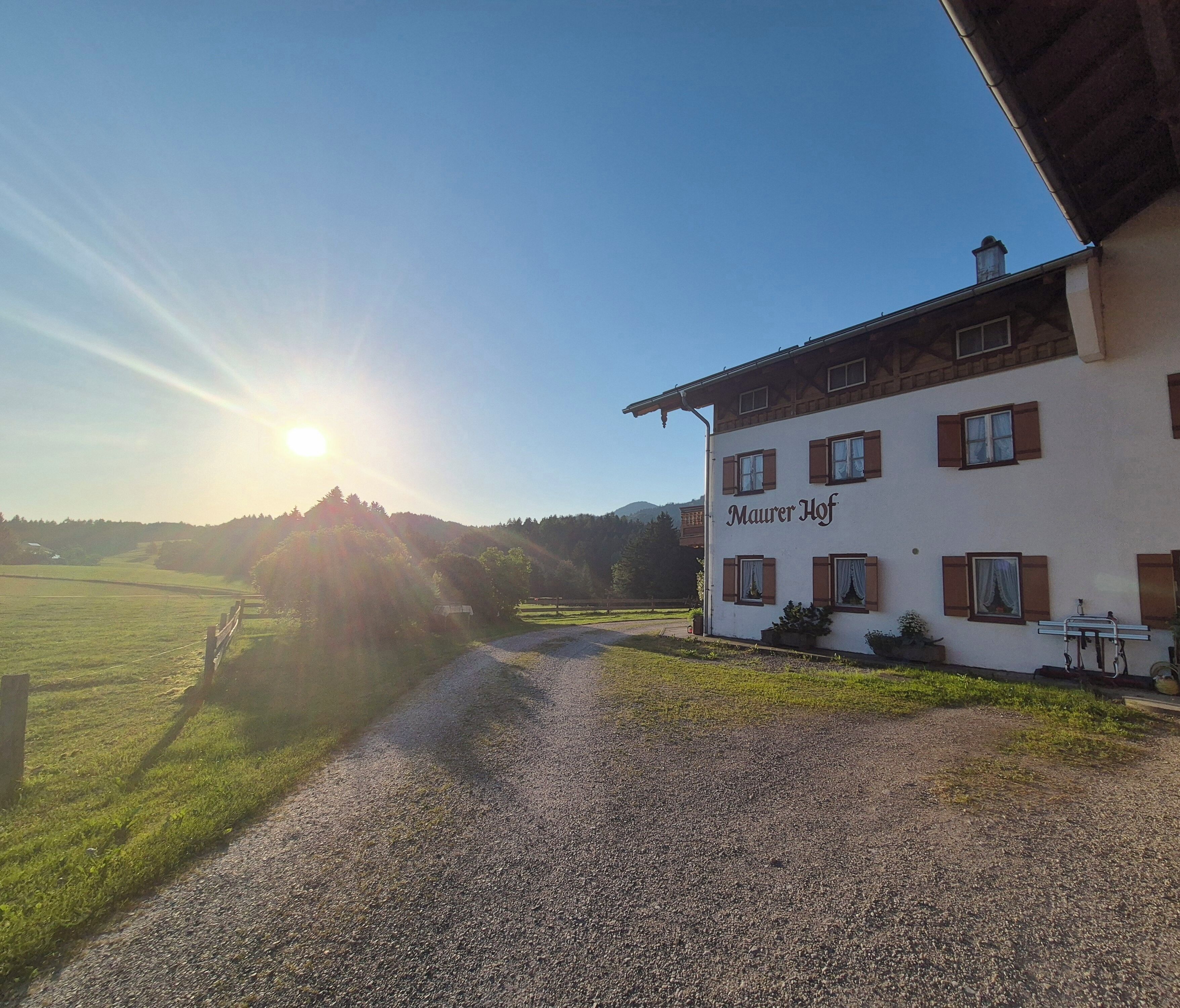 A building with the name 'Maurer Hof' on its facade, a gravel path leading to it, and a clear sky with the sun low on the horizon.