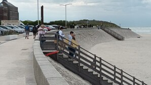 Ein Küstenweg mit Holzgeländer, ein Sandstrand und ein Parkplatz mit Autos.