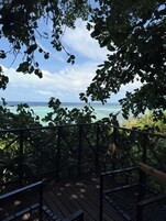 Um deck de madeira com guarda-corpo, com vista para uma praia com águas turquesa cristalinas e céu parcialmente nublado.