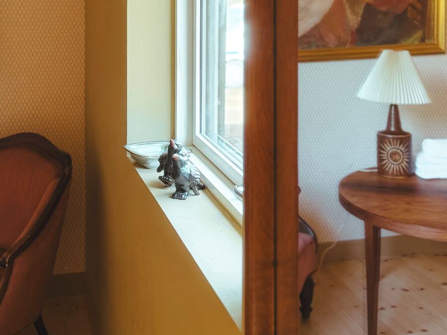 Una habitación con ventana, un cuadro en la pared, una lámpara en una mesita auxiliar y una silla.