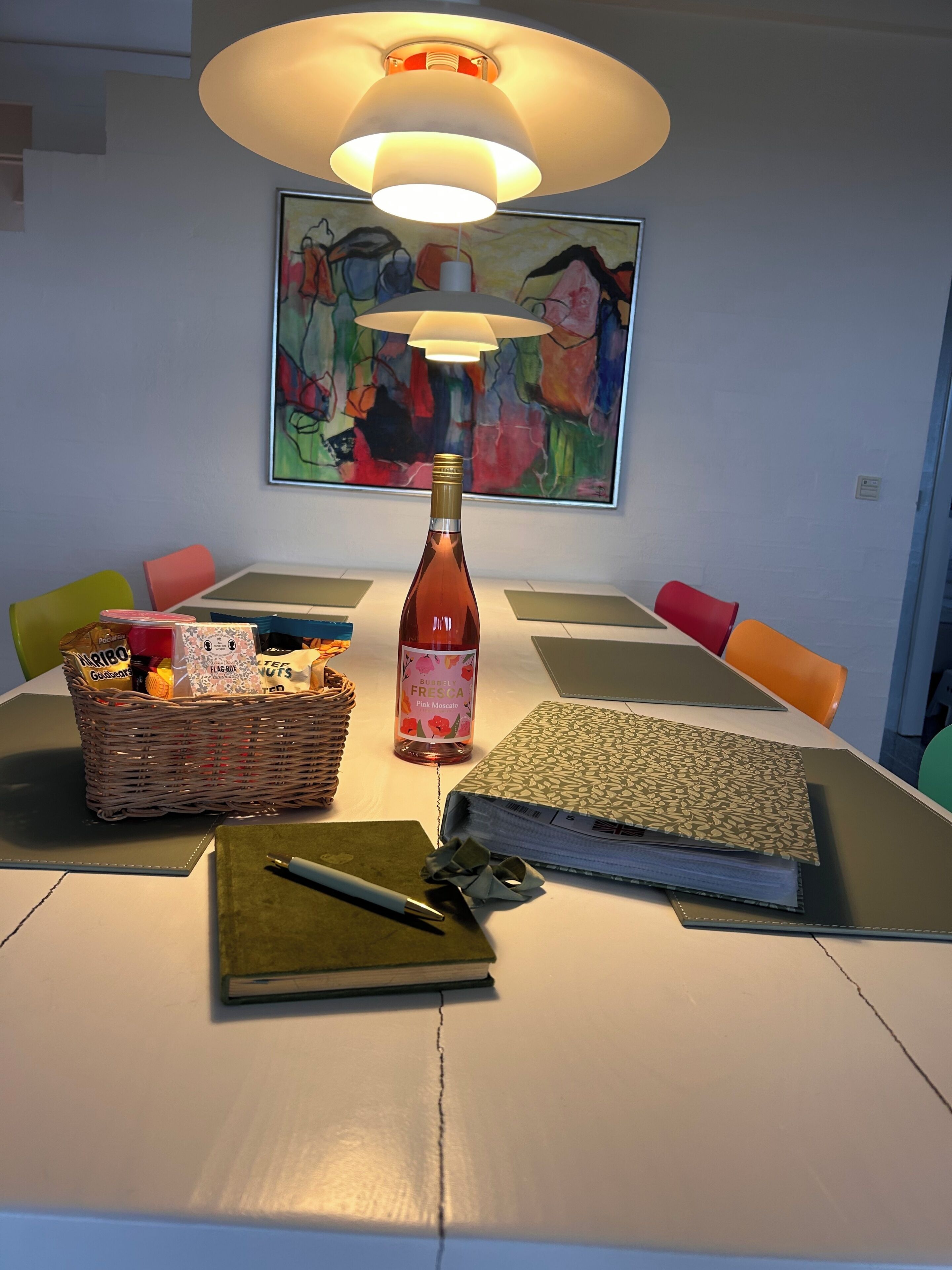 A dining table with a bottle of wine, a basket of snacks, a notebook, and a pen.
