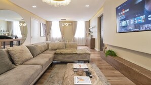 A modern living room with a sectional sofa, a coffee table, and a TV mounted on the wall displaying a program. - Cozy 3 Bedroom Apartmen -Swimming Pool & Gym QQ (Lagos)