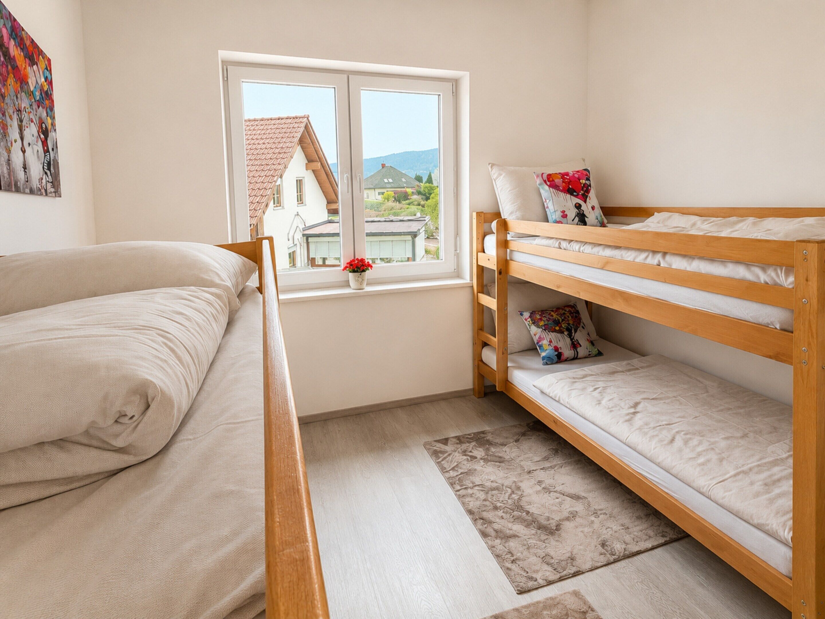 A bedroom with a bunk bed, a single bed, a window with a view of houses, and a painting on the wall.