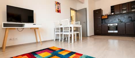 A modern living room with a flat-screen TV, white chairs, a colorful rug, a kitchen with dark cabinets, and a ceiling light.