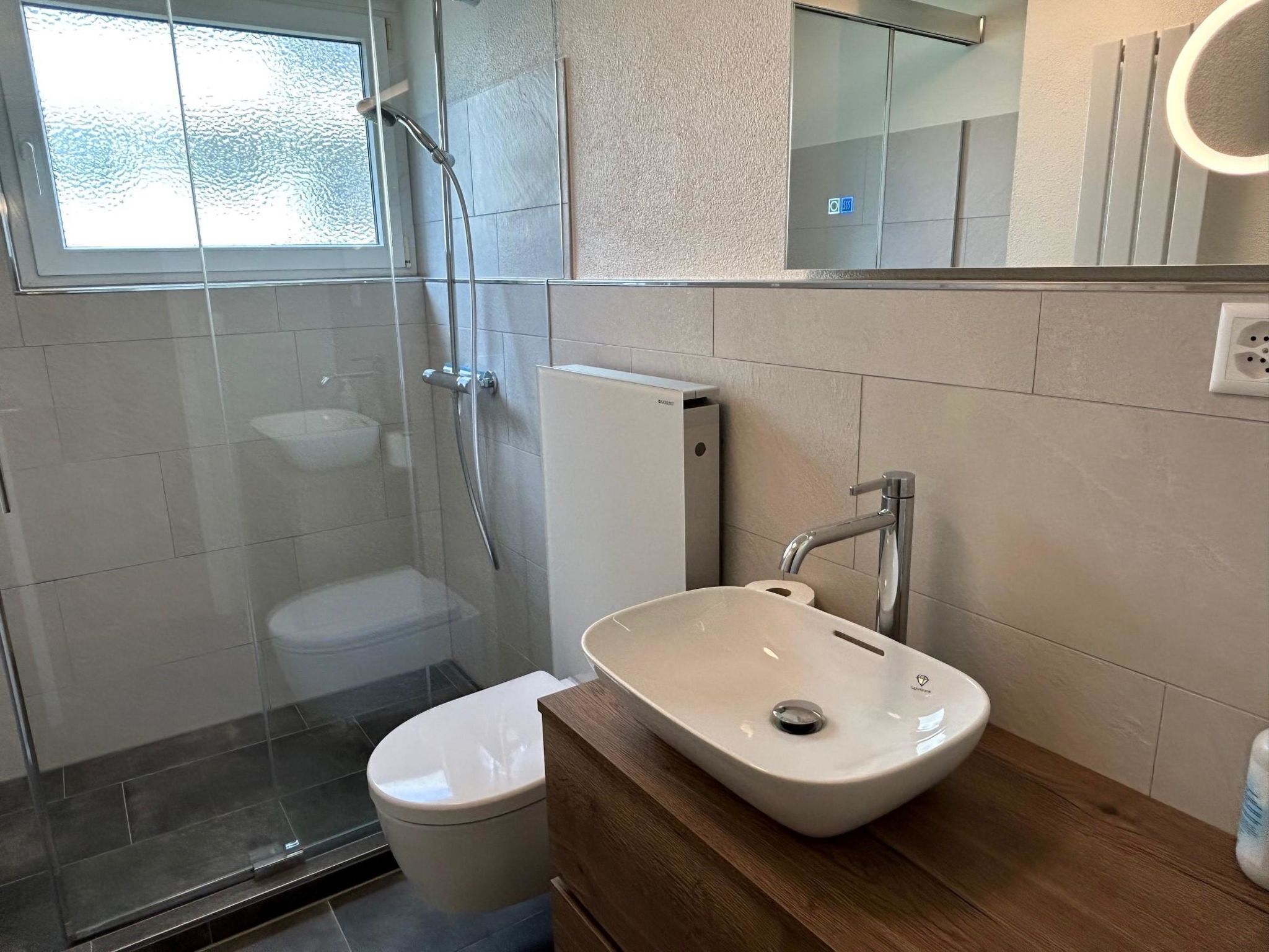 Uma casa de banho moderna com cabine de duche em vidro, sanita branca e lavatório com bancada de madeira.