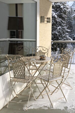 Metallstühle und ein Tisch auf einem Balkon, im Hintergrund verschneite Bäume.