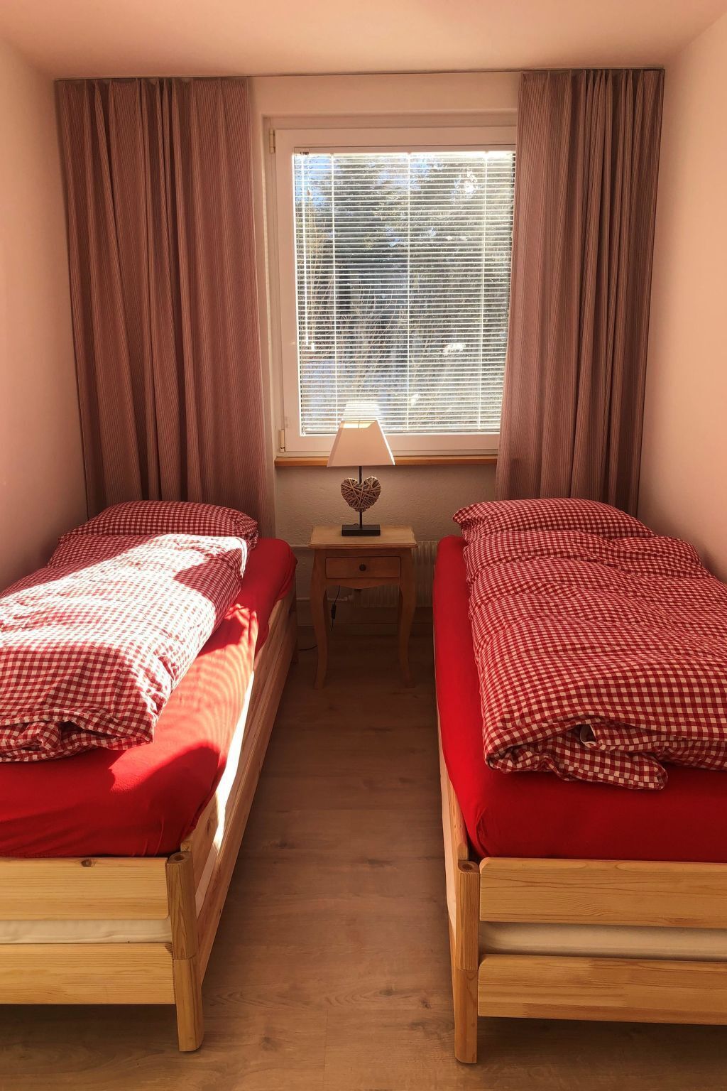 Una habitación con dos camas de madera, una mesita de noche con una lámpara y una ventana con persianas.