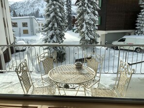 A balcony with a round table and chairs, overlooking a snowy street with buildings and a van. - Alkazar Wg. 11 Appartement/Fewo, Bad, WC, 6-Bettwohnung by Interhome (Arosa)