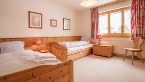 Een slaapkamer met twee houten bedden, een nachtkastje, een stoel, een raam met gordijnen en foto's aan de muur.