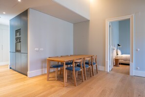 Een moderne keuken met inbouwapparatuur, een eethoek met een houten tafel en blauwe stoelen, en een slaapkamer zichtbaar door een open deur.