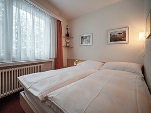 Ein Hotelzimmer mit einem großen Bett, einem Heizkörper, einem fenster mit Vorhängen, einer Vase und zwei gerahmten Bildern an der Wand.
