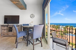 Un balcón con vistas al océano, una zona de comedor con mesa y sillas, un televisor sobre un mueble de madera y un reloj de pared.