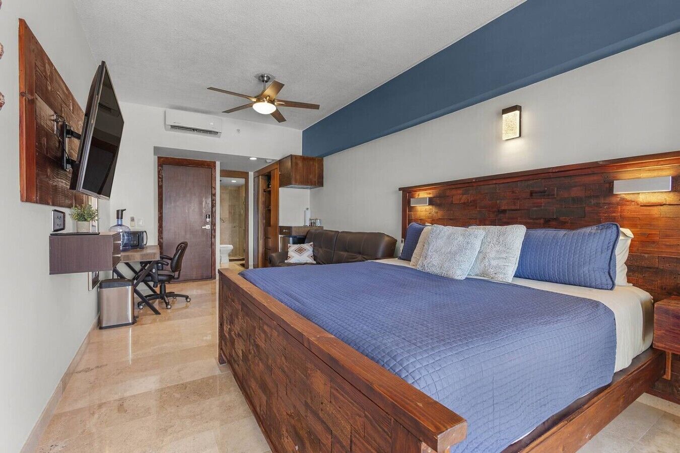A bedroom with a large bed, a desk, a chair, a television, and a ceiling fan.