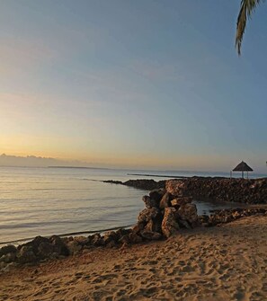 A beach with a rock formation, a thatched umbrella, and a calm sea. - Azure Coast Resort Hotel (Dar es Salaam)