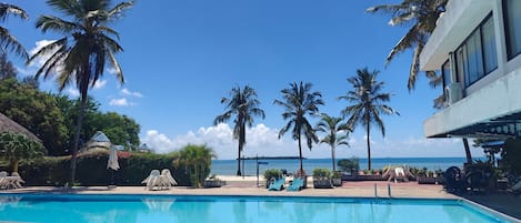 A swimming pool with a dolphin mosaic, surrounded by palm trees and lounge chairs, with a view of the ocean.