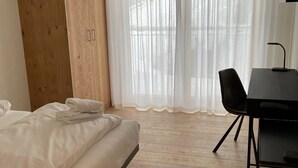 Ein Zimmer mit einem Bett, einem Holzschrank, einem Schreibtisch mit Stuhl und leicht transparenten Vorhängen.