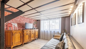 A living room with a sofa, wooden cabinet, television, and a brick wall. - Warm Apartment a/c 2br/6p - Nice (Nice)