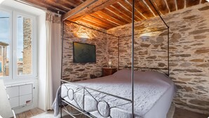Un dormitori de parets de pedra, amb estructura de llit metàl·lica, un piló de pedra i una finestra amb cortines.