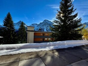 A modern building with a flat roof, surrounded by snow-covered ground and tall evergreen trees, with a mountain range in the background. - Utoring Hohe Promenade 004 Appartement/Fewo, Dusche, WC, 2-Bettwohnung by Interhome (Arosa)