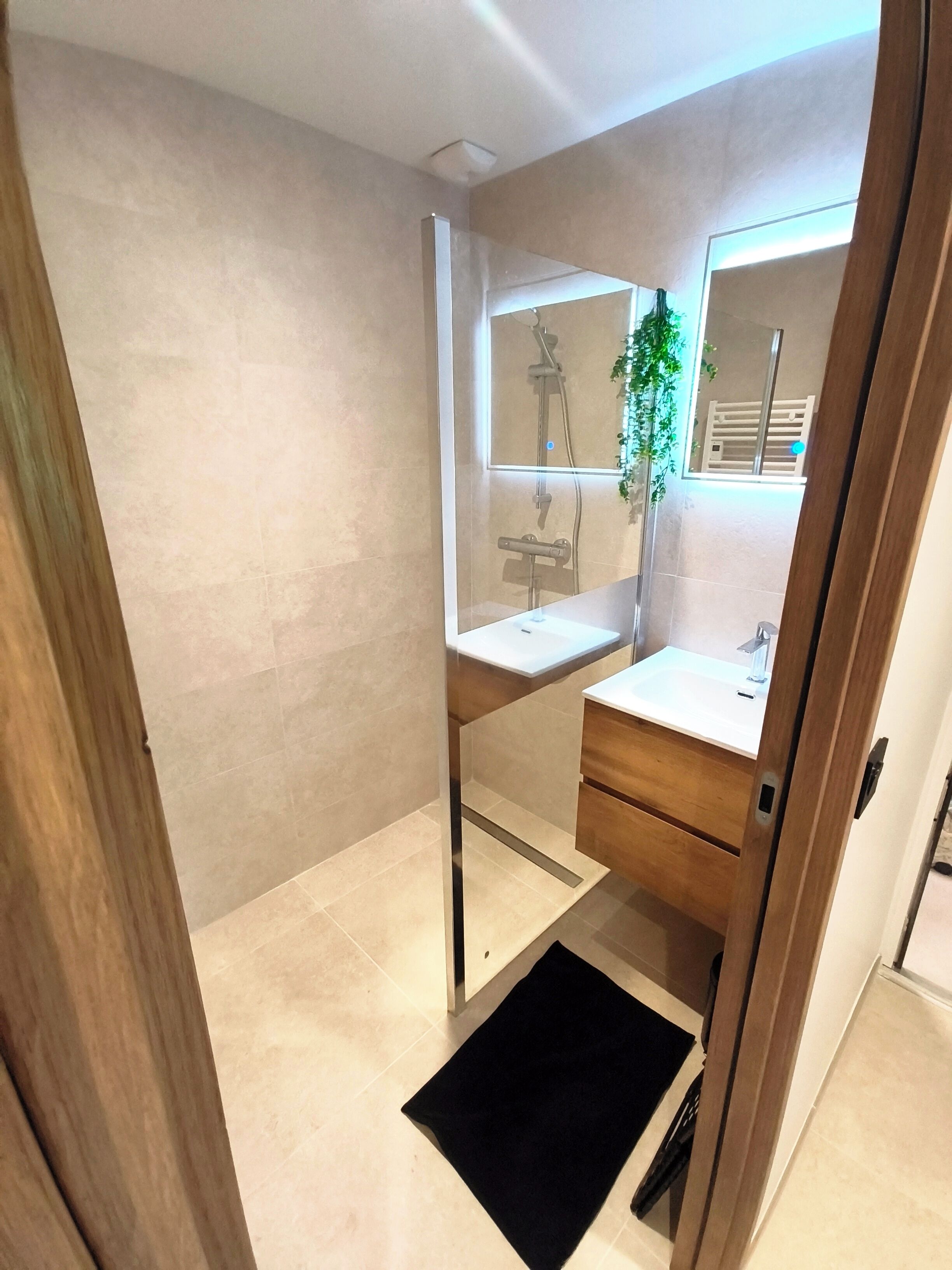 A modern bathroom with a glass shower enclosure, a white sink, a mirror, and a black mat.