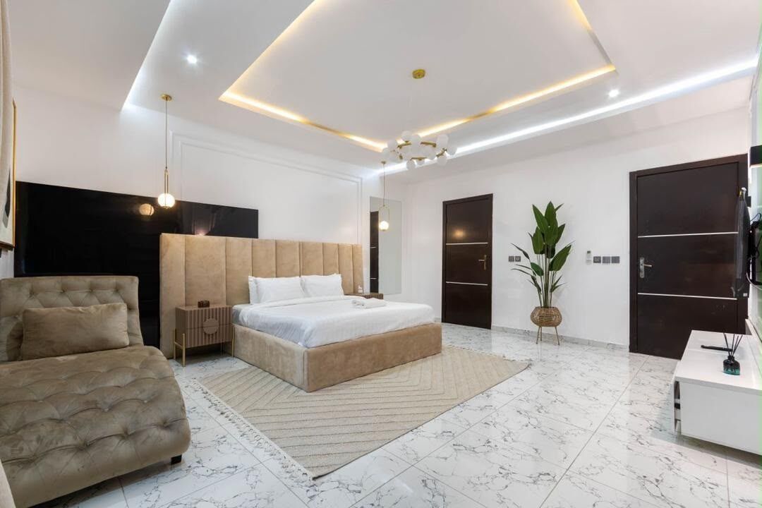 A modern bedroom with a large bed, a sofa, a bedside table, and a potted plant.