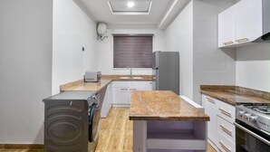 Een kleine keuken met witte kastjes, een granieten aanrecht, een inbouwoven en een raam met zonwering.