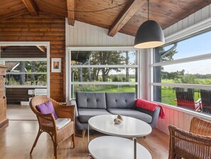 A cozy room with a wooden ceiling, a grey sofa, a wicker chair, a white table, and large windows offering a view of greenery.