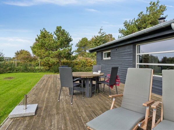 A deck with a table and chairs, a small house with a window, and a grassy yard with trees.