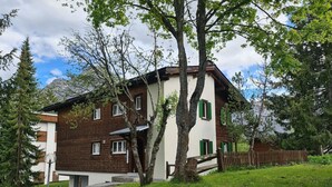 Een gebouw met twee verdiepingen, een bakstenen gevel en groene luiken, omringd door bomen en gazon.