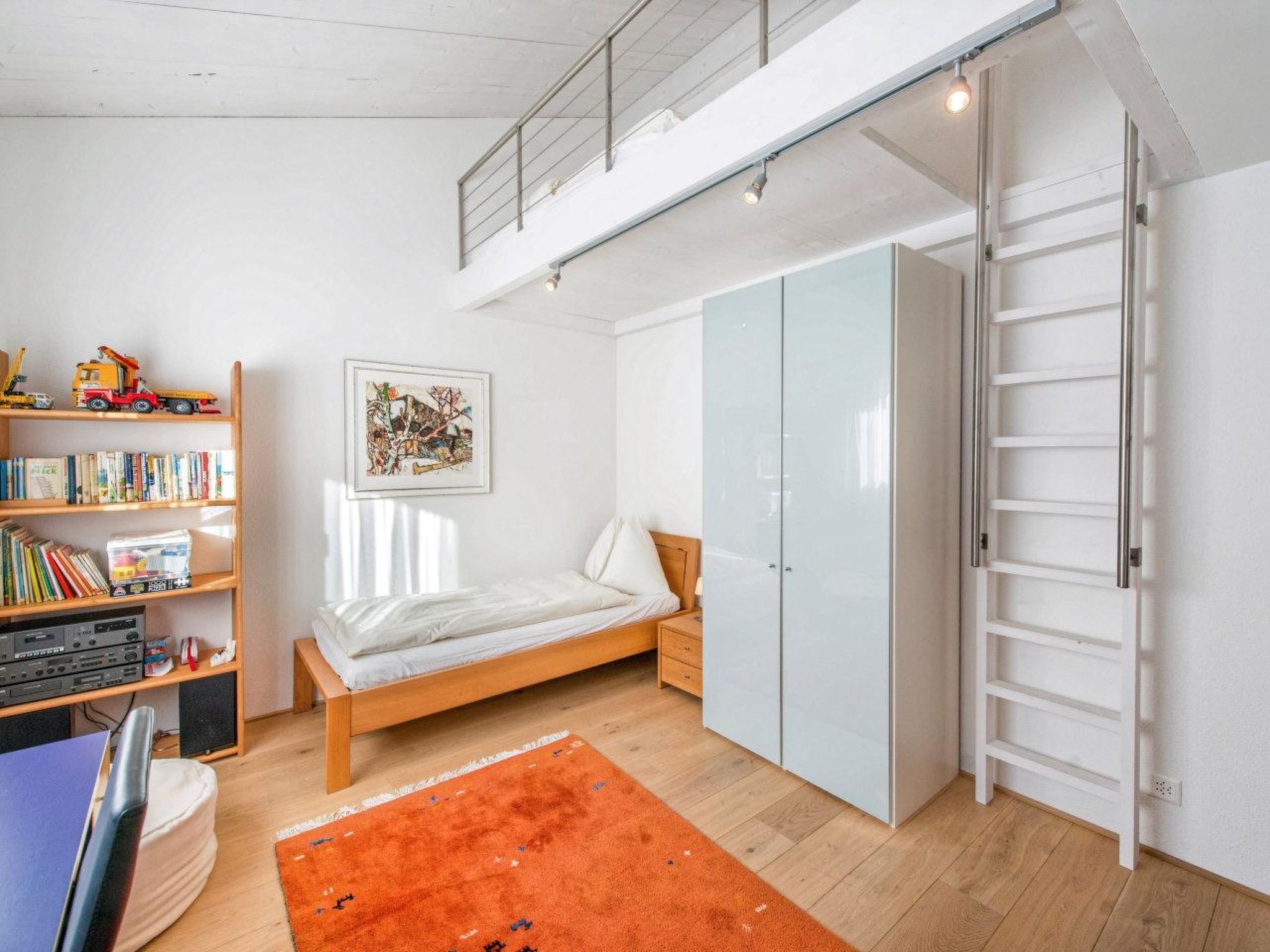 A bedroom with a loft bed, a bookshelf, a wardrobe, and a desk with a computer.